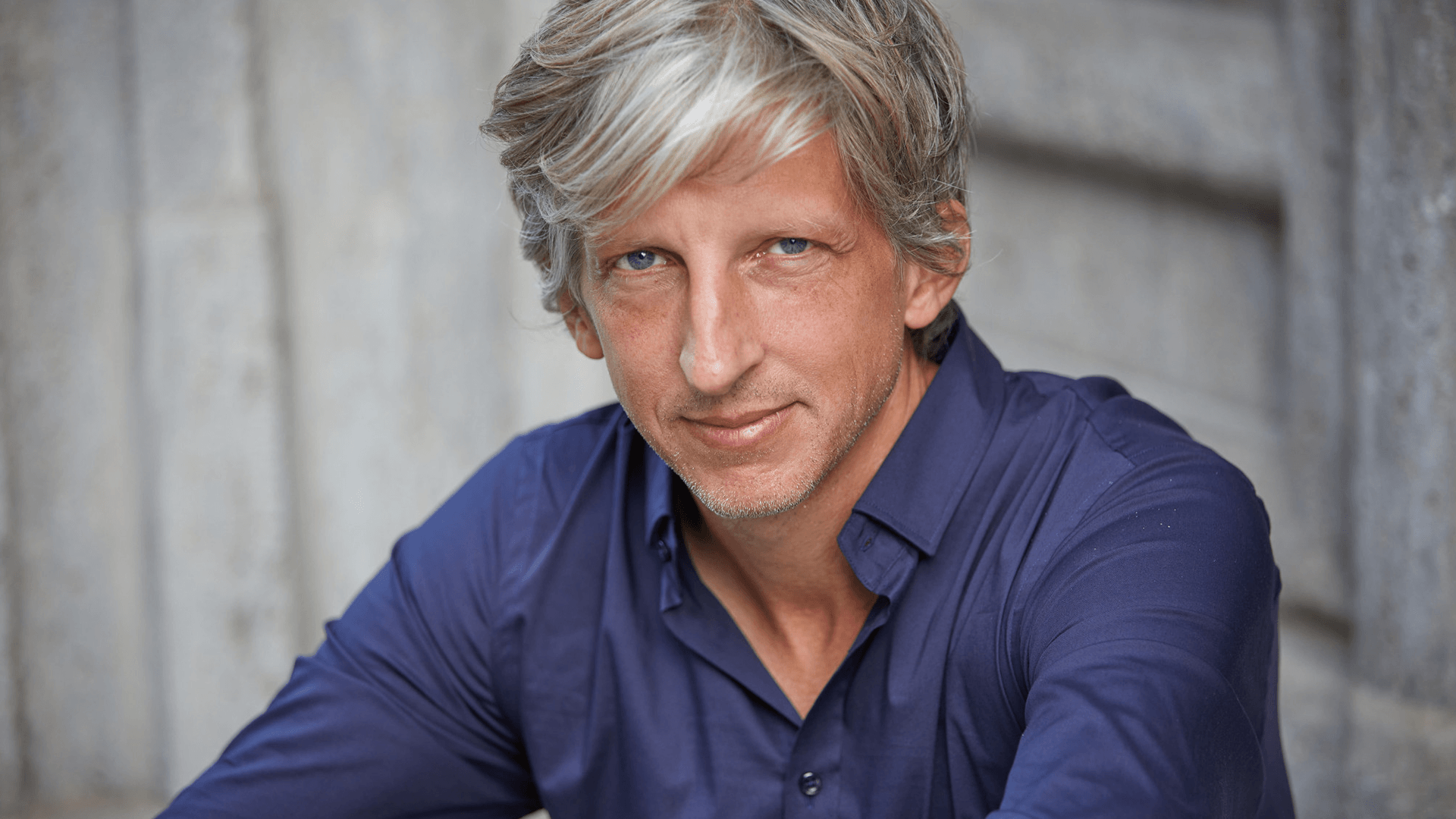 Pressebild William Wahl. William schaut leicht lächelnd in die Kamera und trägt ein blaues Hemd.